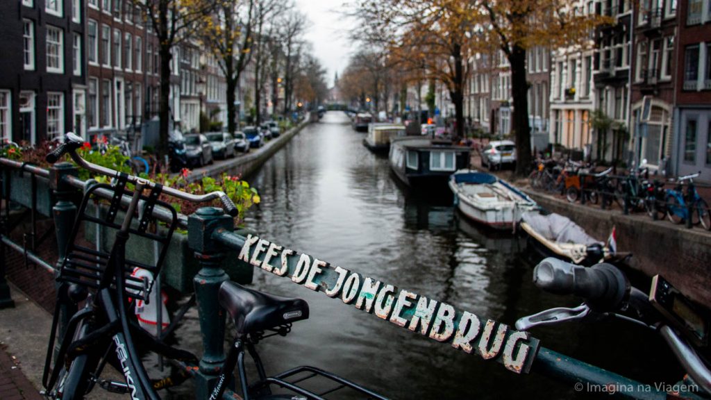 Canal de Amsterdam visto de uma ponte, com bicicletas em primeiro plano e barcos entre casas estreitas nas margens