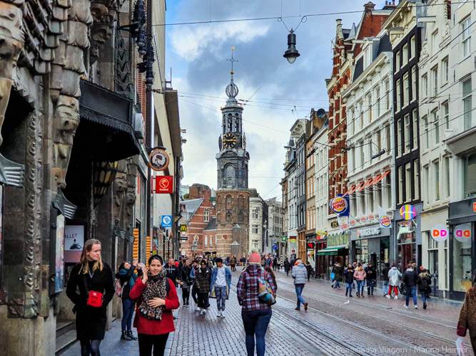 Munttoren ao fundo de uma rua movimentada no centro de Amsterdam, cercada por lojas e pedestres