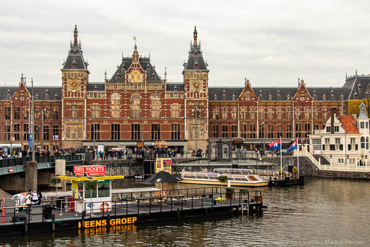 Fachada da Amsterdam Centraal vista do canal, com barcos e plataformas de embarque em primeiro plano